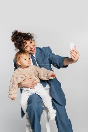 happy businesswoman in blue suit taking selfie with toddler daughter on chair isolated on grey 