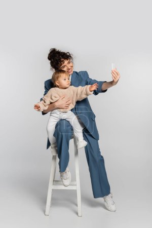 cheerful businesswoman in blue suit taking selfie with toddler daughter on high chair on grey 
