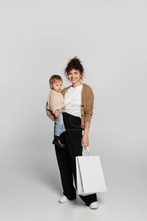 full length of happy mother in casual clothes holding in arms cheerful baby daughter and shopping bag on grey 
