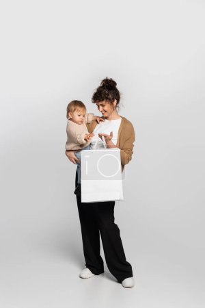 full length of cheerful mother in casual clothes holding in arms daughter and shopping bag on grey 