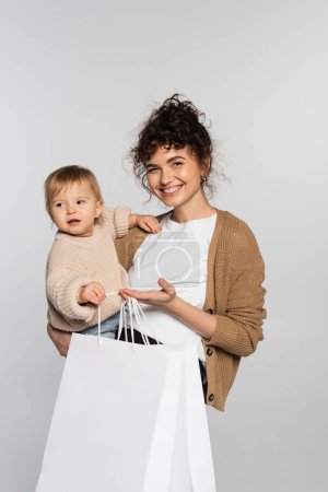 cheerful mother in casual clothes holding in arms daughter and shopping bags isolated on grey 