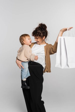 happy mother in casual clothes holding in arms toddler daughter and shopping bags isolated on grey 