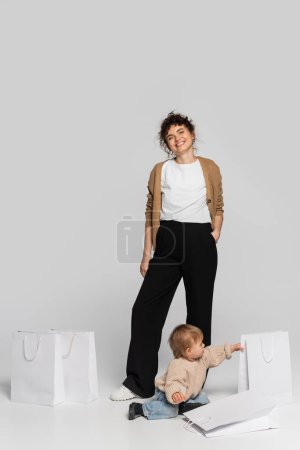 full length of happy woman in casual clothes posing with hand in pocket near toddler daughter and shopping bags on grey 