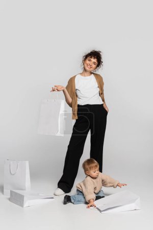 full length of cheerful woman in casual clothes posing with hand in pocket near toddler daughter and shopping bags on grey 