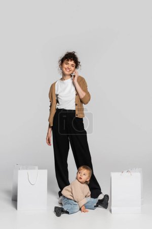 full length of cheerful woman in casual clothes talking on smartphone near toddler daughter and shopping bags on grey 