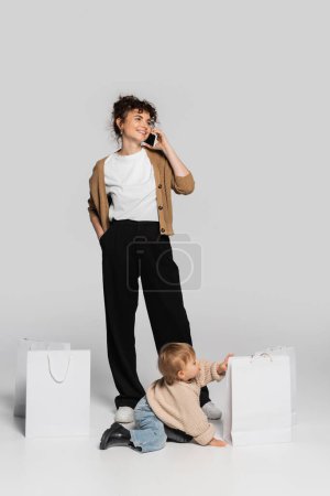 full length of happy woman in casual clothes talking on smartphone near toddler baby and shopping bags on grey 