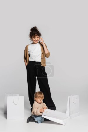 full length of cheerful woman talking on smartphone near toddler girl and shopping bags on grey 