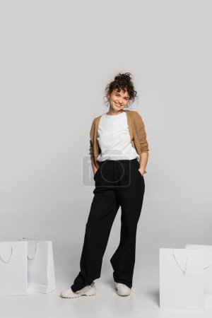 full length of cheerful woman in casual clothes standing with hands in pockets near shopping bags on grey 