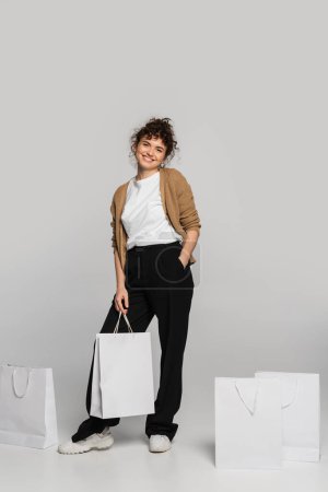 full length of cheerful woman in casual clothes standing with hand in pocket near shopping bags on grey 