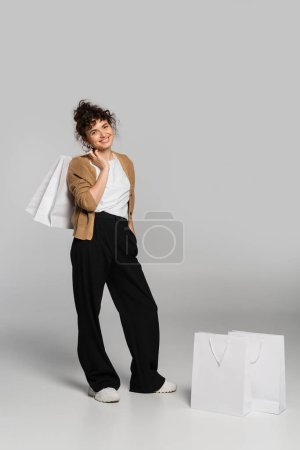 full length of cheerful woman in casual clothes standing with hand in pocket and holding shopping bag on grey 