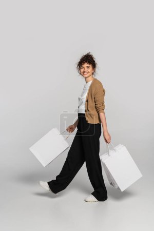 Téléchargez les photos : Full length of cheerful woman in casual clothes walking with shopping bags on grey - en image libre de droit