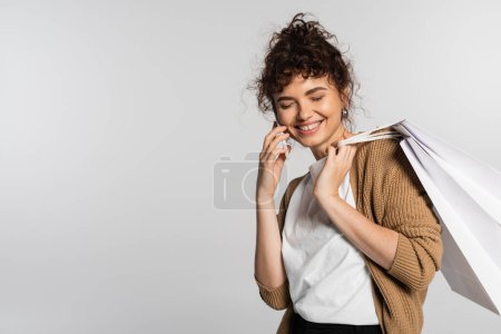 cheerful woman in casual clothes talking on smartphone and holding shopping bags isolated on grey 