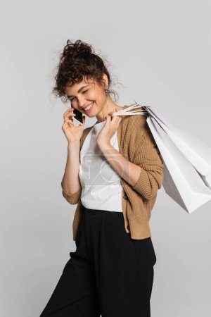 Téléchargez les photos : Positive woman in casual clothes talking on smartphone and holding shopping bags isolated on grey - en image libre de droit