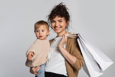 cheerful mother in casual clothes holding in arms toddler daughter and shopping bags isolated on grey 