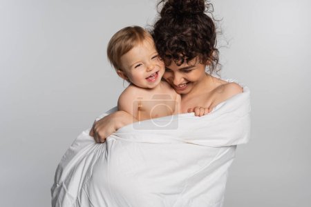 cheerful mother and toddler baby girl covered in duvet isolated on grey 