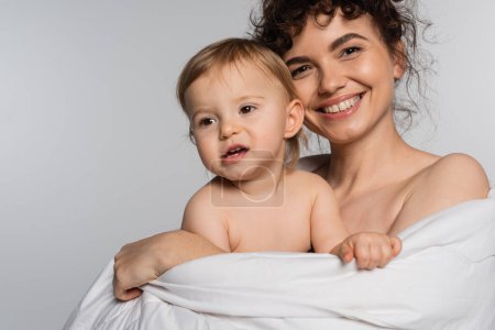 smiling mother and toddler baby girl covered in duvet isolated on grey 
