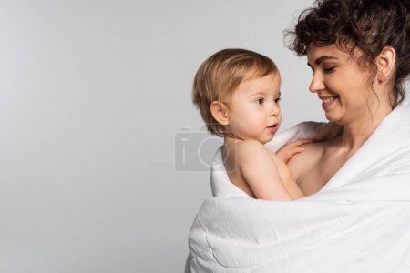 happy mother looking at toddler baby girl covered in duvet isolated on grey 