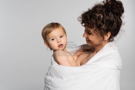 pleased mother and toddler baby girl covered in duvet isolated on grey 
