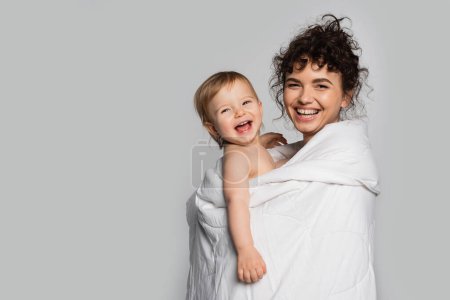 happy mother and positive toddler daughter covered in duvet isolated on grey 