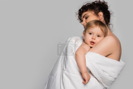 curly mother hugging toddler daughter covered in white duvet isolated on grey 