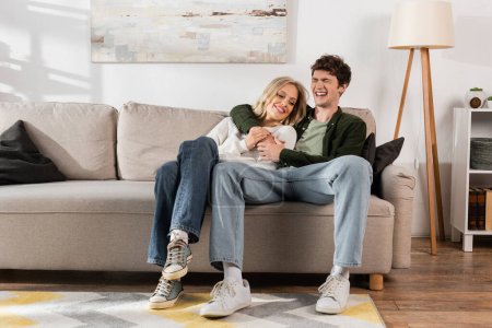 full length of cheerful and curly man hugging happy blonde girlfriend in living room 