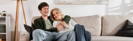 cheerful and curly man hugging happy blonde girlfriend in living room, banner 