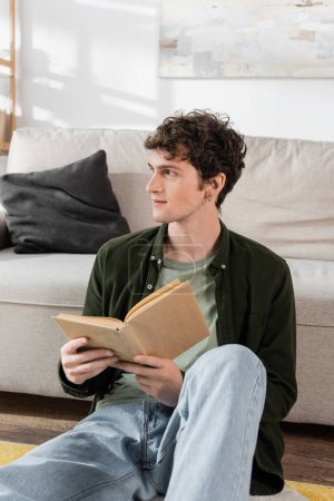 intelligent man with curly hair holding book while looking away in living room 