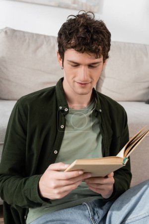 intelligent man with curly hair reading book in living room 