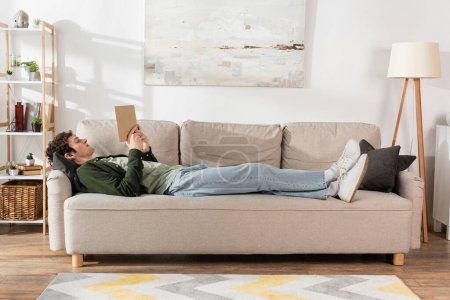 Foto de Full length of young man with curly hair reading book while lying on couch in living room - Imagen libre de derechos