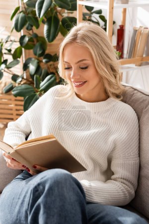 happy blonde woman in white sweater and jeans reading book in living room 