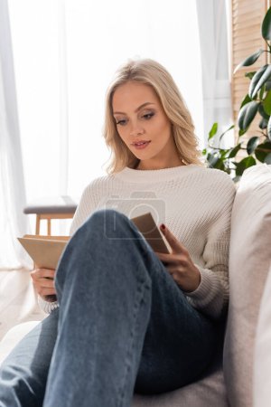 pretty blonde woman in white sweater and jeans reading book in living room 