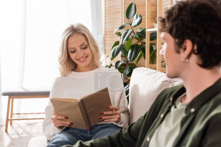 happy blonde woman in white sweater and jeans reading book near boyfriend on blurred foreground 
