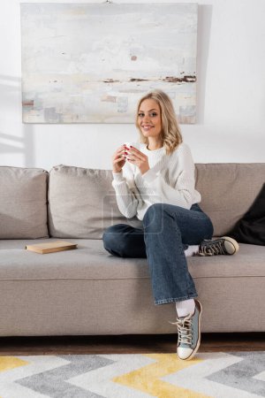 full length of cheerful young woman in white sweater holding cup of coffee at home 