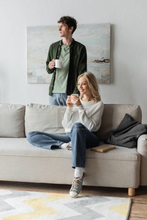 Photo for Curly young man and happy blonde woman holding cups with coffee in living room - Royalty Free Image