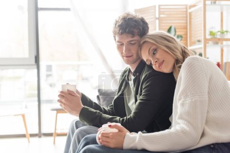 Photo for Blonde woman in sweater holding cup of coffee and leaning on boyfriend in living room - Royalty Free Image