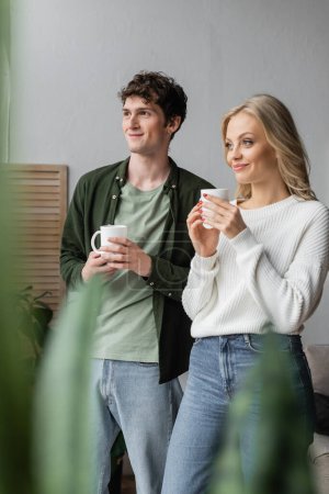 young couple in casual clothes smiling and holding cups of tea near plant on blurred foreground 