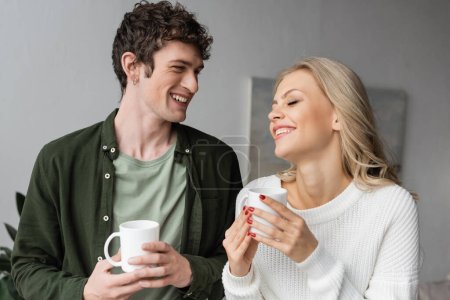 cheerful young couple in casual clothes smiling and holding cups of tea at home 
