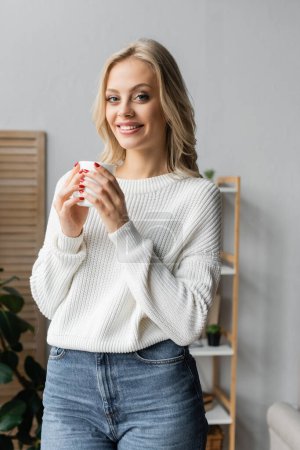 cheerful blonde woman in white sweater holding cup of tea at home 