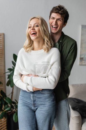excited man with curly hair hugging girlfriend and laughing at home 