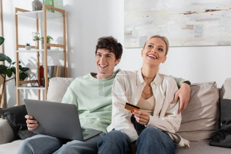 cheerful woman holding credit card near boyfriend with laptop doing online shopping 