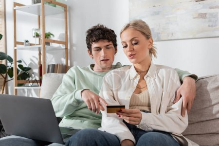 Téléchargez les photos : Curly man hugging blonde girlfriend and pointing at credit card near laptop - en image libre de droit