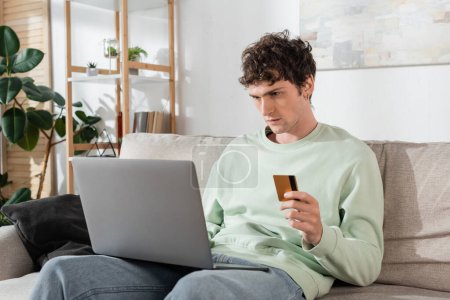 curly young man holding credit card while doing online shopping on laptop 