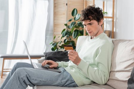 focused young man holding credit card while doing online shopping on laptop 