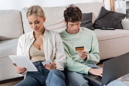 young man and woman holding gadgets and credit cards while doing online shopping 