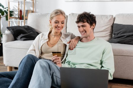 Téléchargez les photos : Cheerful man holding credit card while using laptop near joyful girlfriend - en image libre de droit