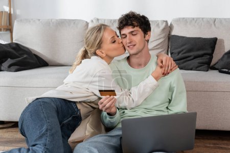 Foto de Happy blonde woman kissing cheek of boyfriend with credit card sitting near laptop - Imagen libre de derechos