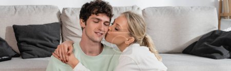 happy blonde woman kissing cheek of boyfriend in living room, banner