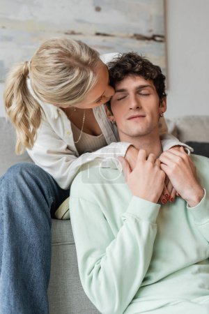 young blonde woman embracing and kissing cheek of curly man in living room 