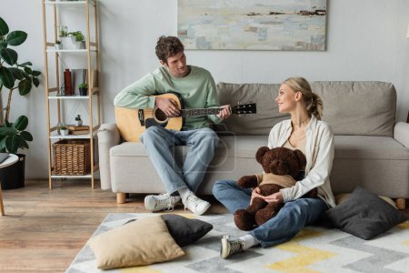 curly man sitting on couch and playing acoustic guitar near happy woman with teddy bear 