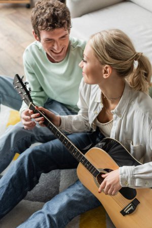 high angle view of cheerful blonde woman playing acoustic guitar near curly boyfriend 